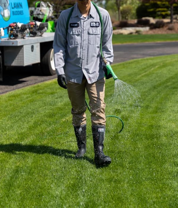 A man wearing a gray shirt and black boots waters a patch of grass with a garden hose.
