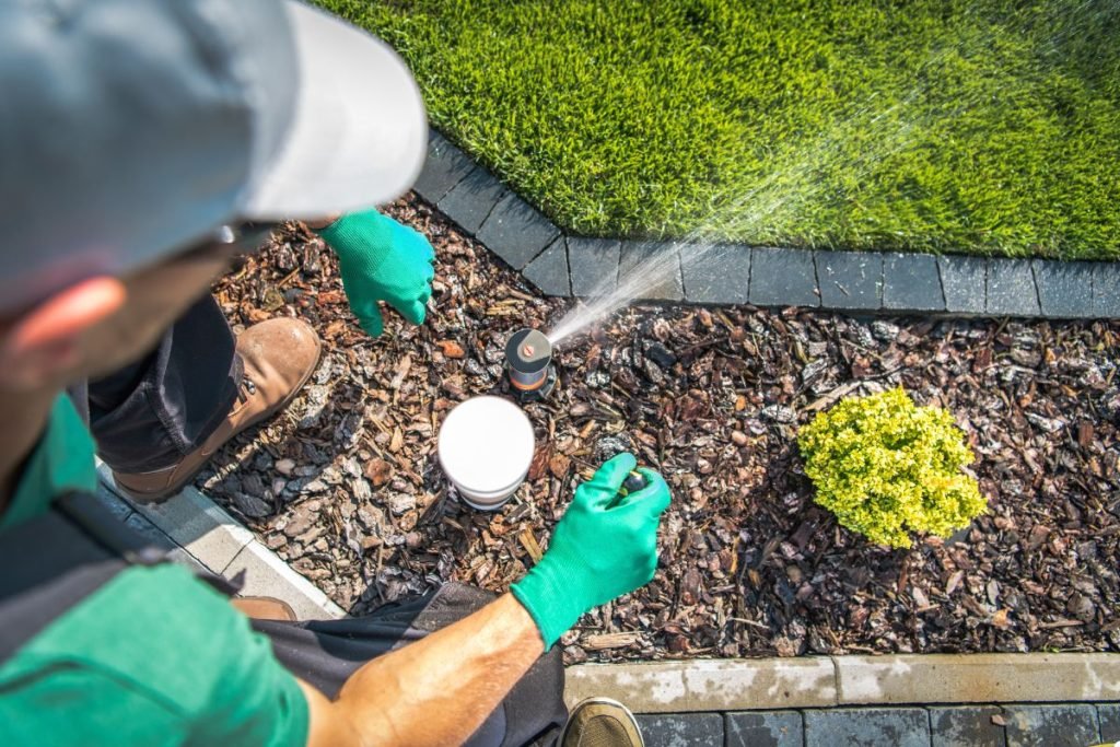 A man in a green shirt and gloves waters plants in a garden, nurturing them with care and attention.